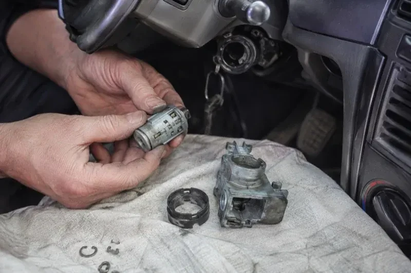 Hands holding a removed ignition cylinder beside a car steering column, with the housing and small parts on a cloth. — Ignition Lock Repair, Kawartha Lakes